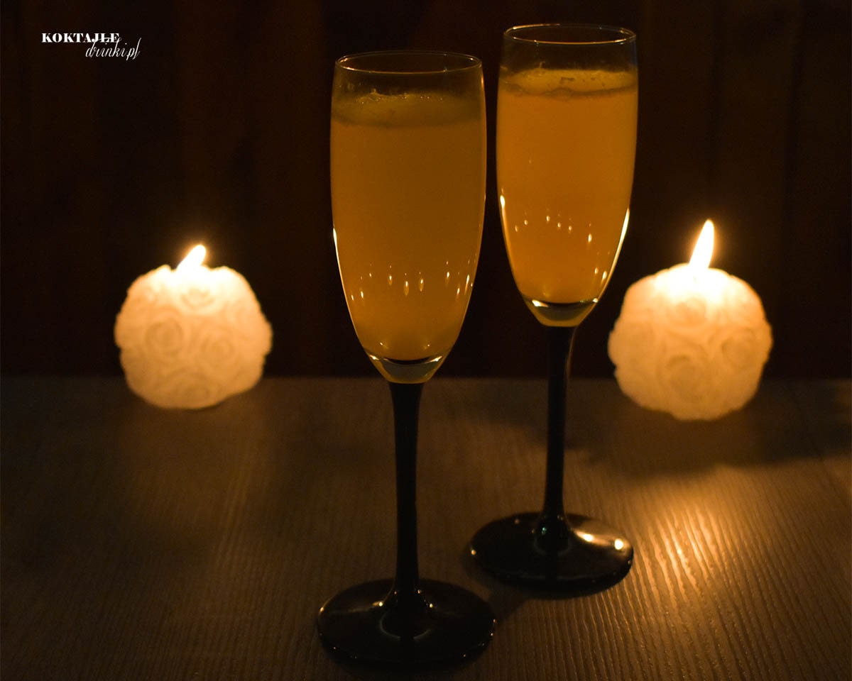 Dwa kieliszki drinka Mimosa, Mimoza z szampanem Prosecco, o pomarańczowej barwie w otoczeniu świec, przy zmroku.