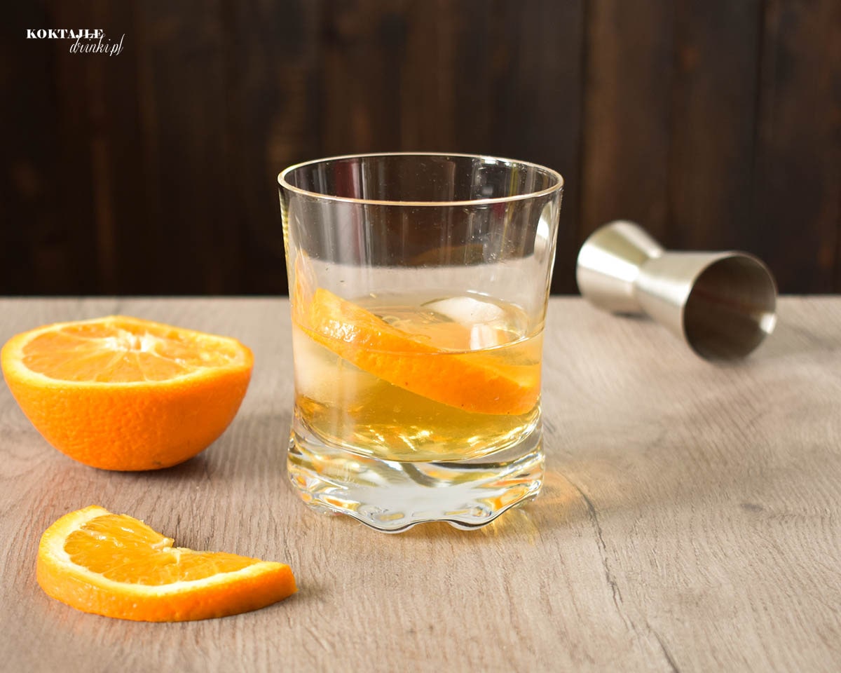 Widok na old fashioned od frontu, złocisty drink w otoczeniu kawałków pomarańczy.
