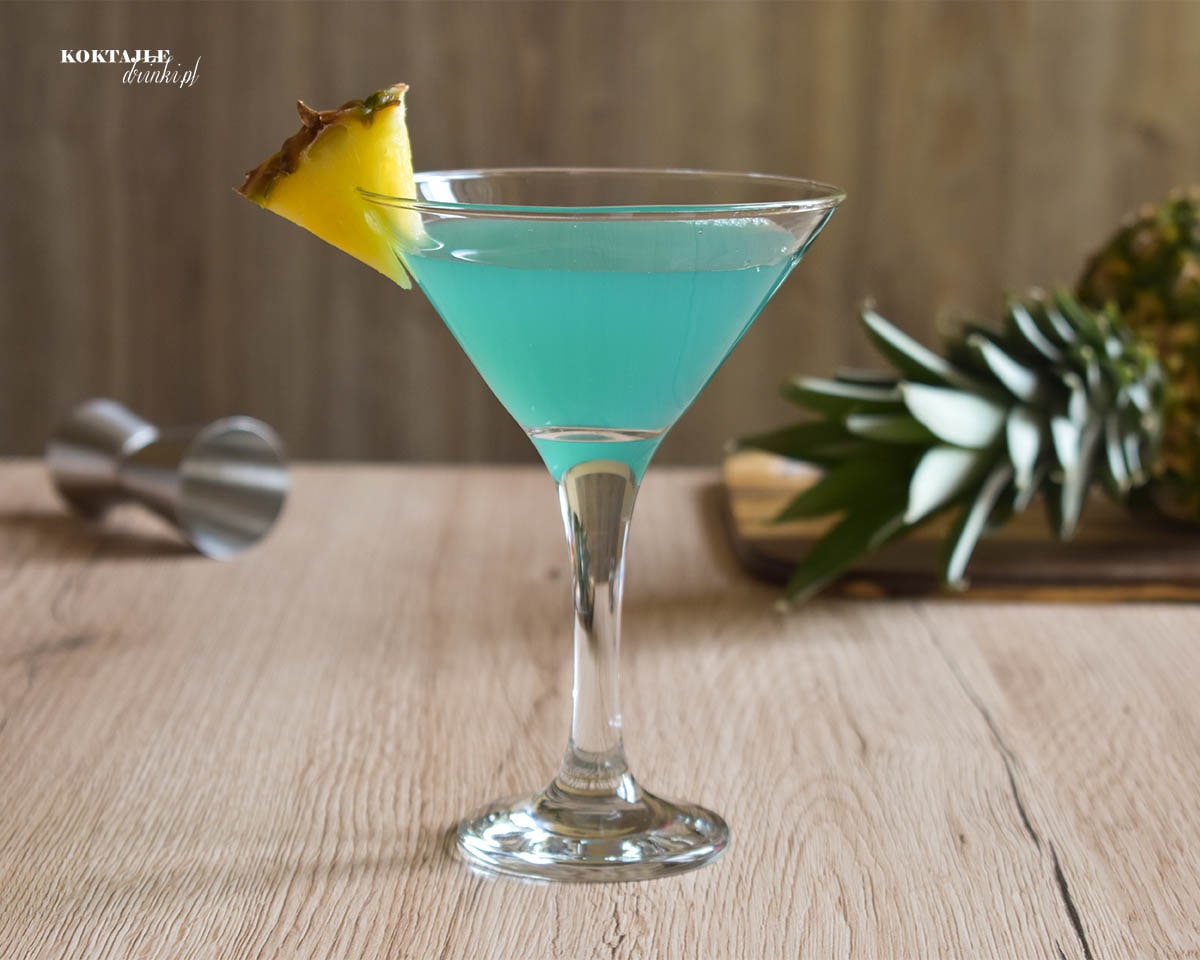 Drink Rajski Ananas z malibu o barwie zielono niebieskiej w kieliszku przyozdobionym kawałkiem ananasa.