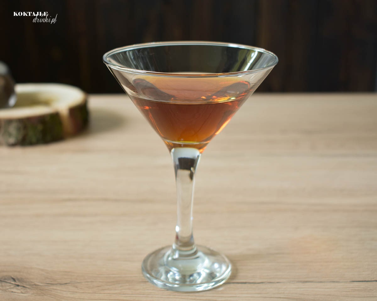 Drink z whisky Revolver o brązowej barwie w kieliszku martini, w tle widoczne kawałek drewna i kieliszek.