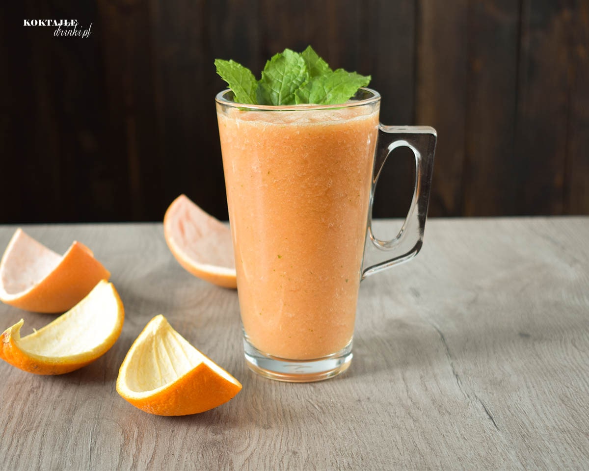 Koktajl owocowy smoothie o barwie pomarańczowej w szklance przyozdobionej miętą, obok leżą skórki pomarańczy oraz grejpfruta.