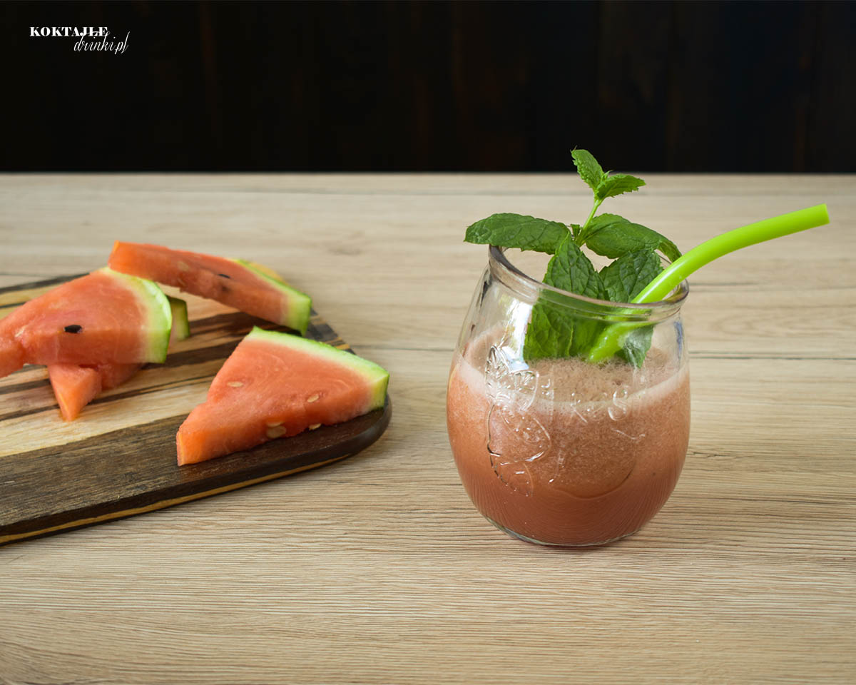 Koktajl owocowy smoothie o barwie jasno czerwonej w otoczeniu kawałków arbuza.